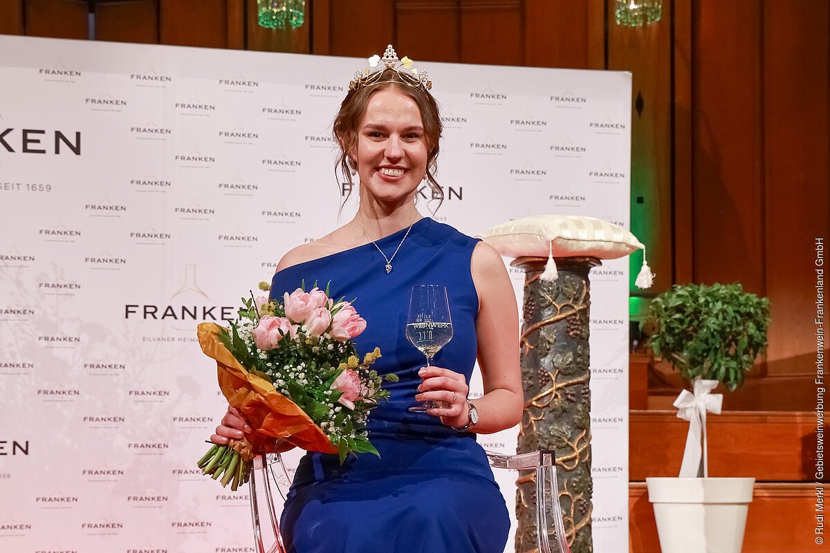 Angelina Seiler heißt die 68. Fränkische Weinkönigin Frau mit Krone, blauem Kleid, Blumenstrauß und Weinglas vor Franken-Werbung sitzend.
