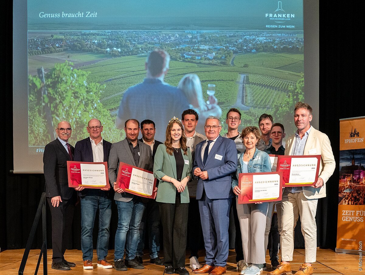 Gruppenfoto von elf Personen auf Bühne mit Urkunden vor Leinwand mit Weinberg-Motiv und Schrift „Genuss braucht Zeit“.