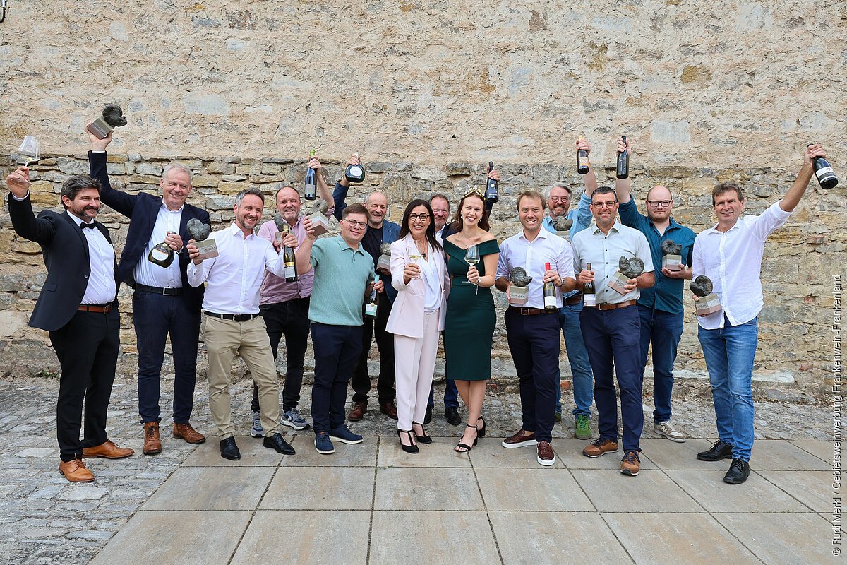 Die Sieger 2026 beim Wettbewerb "Best of Franken" Gruppenfoto von 15 Personen vor einer Steinmauer, die Pokale und Weinflaschen hochhalten.